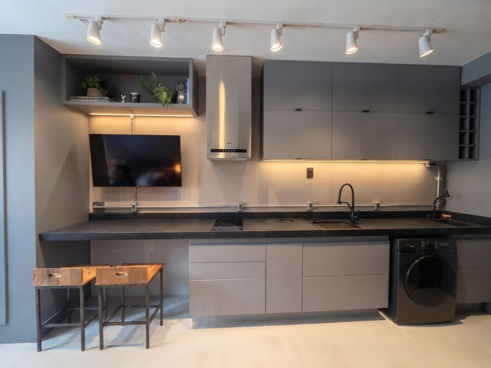 Cozinha linear do Estúdio Brooklin: bancada contínua em pedra preta com cooktop, cuba e banquetas de madeira, TV na parede e lavadora integrada
